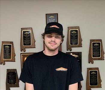 Photo of male employee smiling in black SERVPRO hat and shirt in front of awards hanging on wall