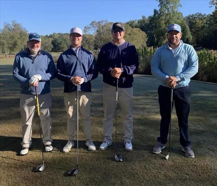 Photo of four men standing with golf clubs at a tournament with trees in background